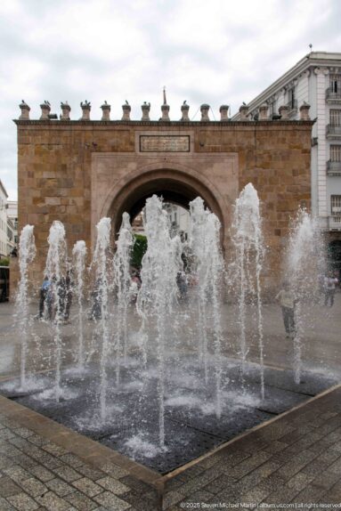 Wide view of Bab el Bhar behind a rectangular fountain basin with multiple vertical water jets.