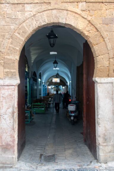 Vaulted Tunis medina market passage with stalls, benches, pedestrians, hanging lamps, and a parked scooter.