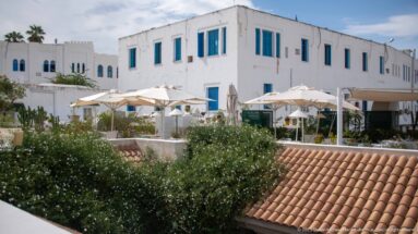White rooftop hotel or guesthouse with blue shutters, café umbrellas, greenery, and terracotta roof tiles.