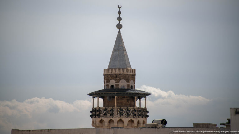 Close upper view of a historic Tunis minaret with arched openings, projecting gallery, pyramidal roof, and crescent finial.