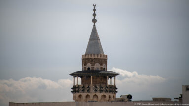Close upper view of a historic Tunis minaret with arched openings, projecting gallery, pyramidal roof, and crescent finial.