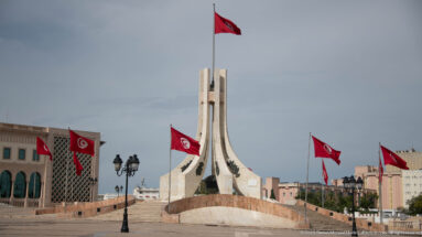 Wide view of the pale Kasbah Square monument surrounded by Tunisian flags, lamps, and government buildings.
