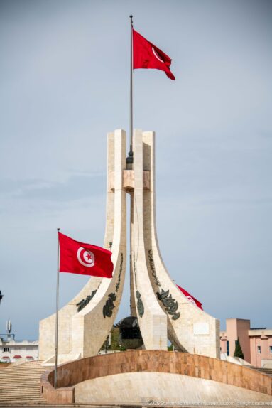 Frontal close view of the Kasbah Square monument with multiple red Tunisian flags and dark decorative motifs.
