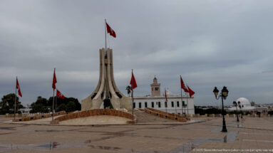 Kasbah Square monument on a wet plaza with Tunisian flags, lamps, white buildings, a minaret, and dome.