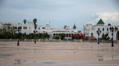 Broad paved Kasbah Square facing white civic buildings, Tunisian flags, palms, towers, domes, and a green roof.