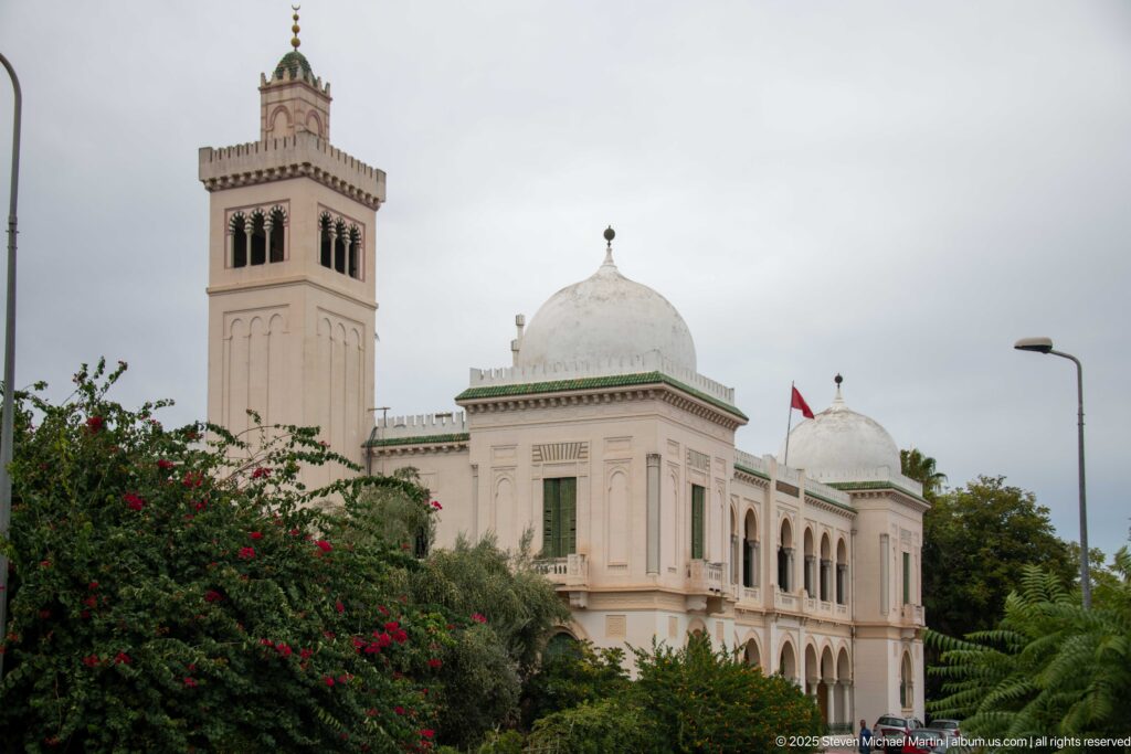 White Tunis civic complex with square tower, domed pavilions, arcaded balconies, greenery, and Tunisian flag.