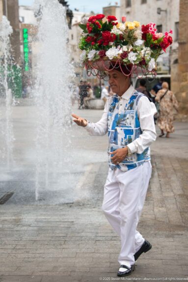 Street flower seller in white clothing carrying a large round display of red, white, and yellow flowers.