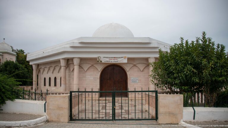 Small pale stone domed building with arched wooden door, narrow windows, bilingual sign, and iron gate.