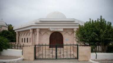 Small pale stone domed building with arched wooden door, narrow windows, bilingual sign, and iron gate.