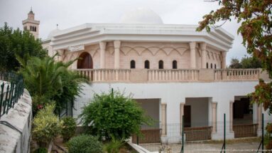 Side view of a pale domed pavilion with pointed arches, columns, balustrade, palms, and garden vegetation.