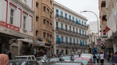 Busy Tunis street with cars, pedestrians, shops, Arabic signs, and a white building with blue shutters and iron balconies.