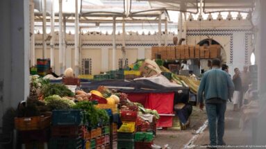 Covered Tunis produce market with stacked vegetable crates, shoppers, stalls, and a high metal-framed roof.
