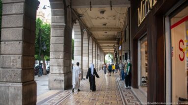 Covered Tunis sidewalk with stone arcades, patterned pavement, pedestrians, mannequins, and shopfronts.