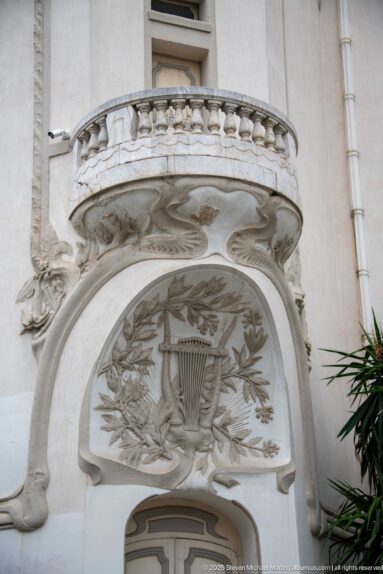 Carved lyre and laurel relief beneath an ornate projecting balcony on a white historic façade in Tunis.