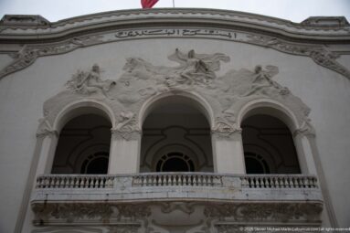 Upper Municipal Theatre of Tunis façade with three arches, balustrade, sculptural reliefs, and Arabic inscription.
