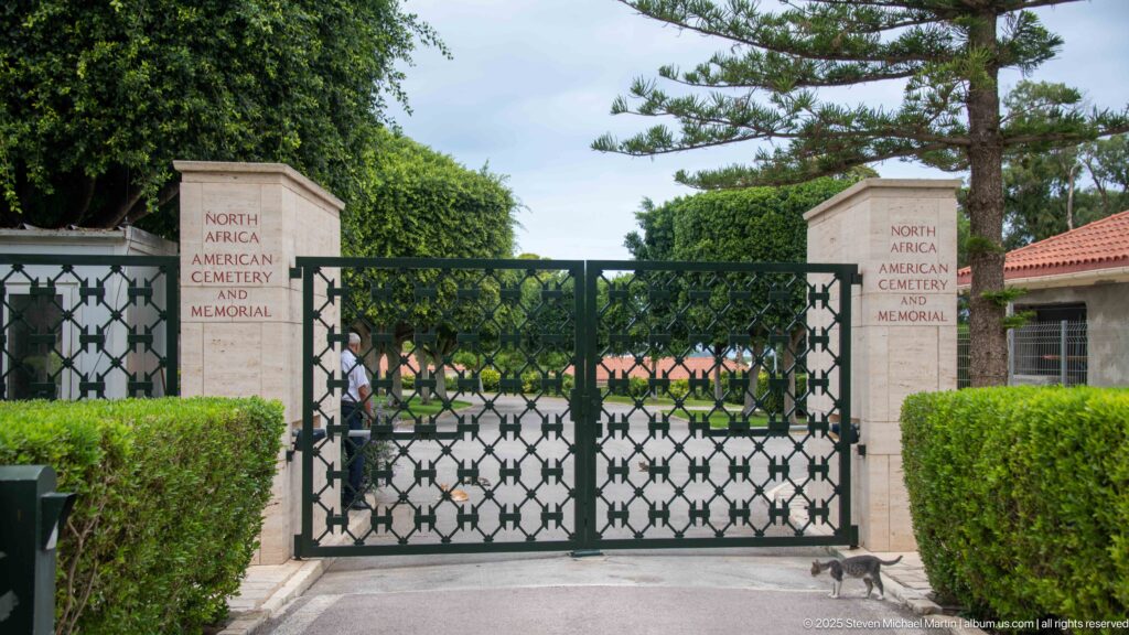Stone entrance pillars and a dark metal gate open into clipped hedges and formal cemetery grounds.