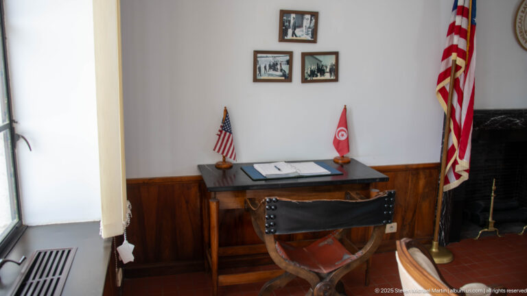 Desk with guest book beneath historic photographs, flanked by small U.S. and Tunisian flags and a larger American flag.