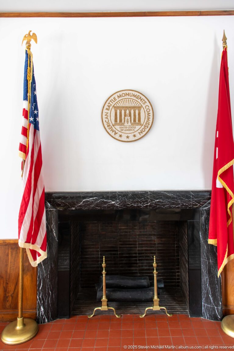 Dark marble fireplace beneath the ABMC seal, flanked by an American flag and a red ceremonial flag.