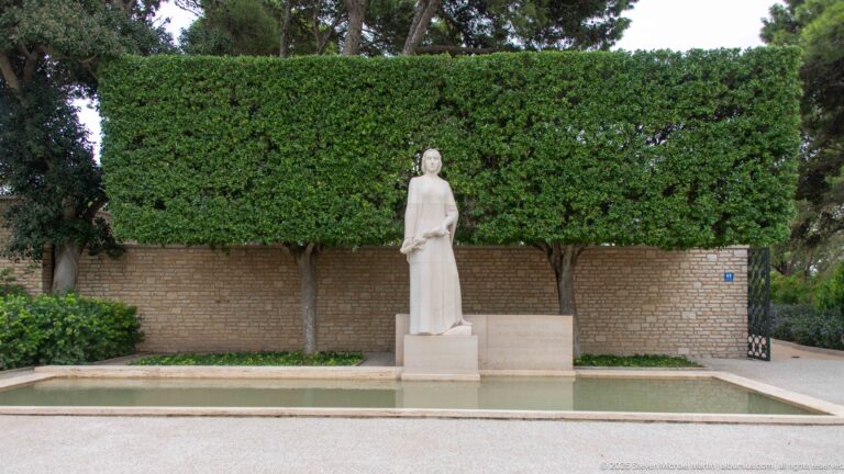 Pale stone standing female figure beside a shallow reflecting pool, backed by clipped hedge and masonry wall.