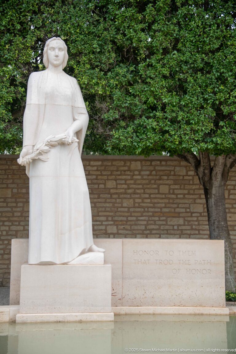 Rear view of a draped female stone figure on a pedestal beside an inscription wall and shallow pool.