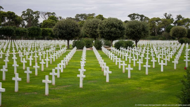 Rows of white Latin-cross grave markers extend across a green lawn beneath mature trees.