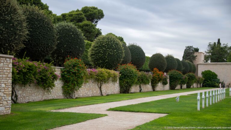Curving pale path beside a stone wall, flowering shrubs, clipped trees, and a few white grave markers.