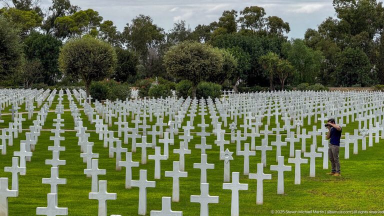 Visitor stands beside extensive rows of white cross-shaped grave markers on a green lawn.