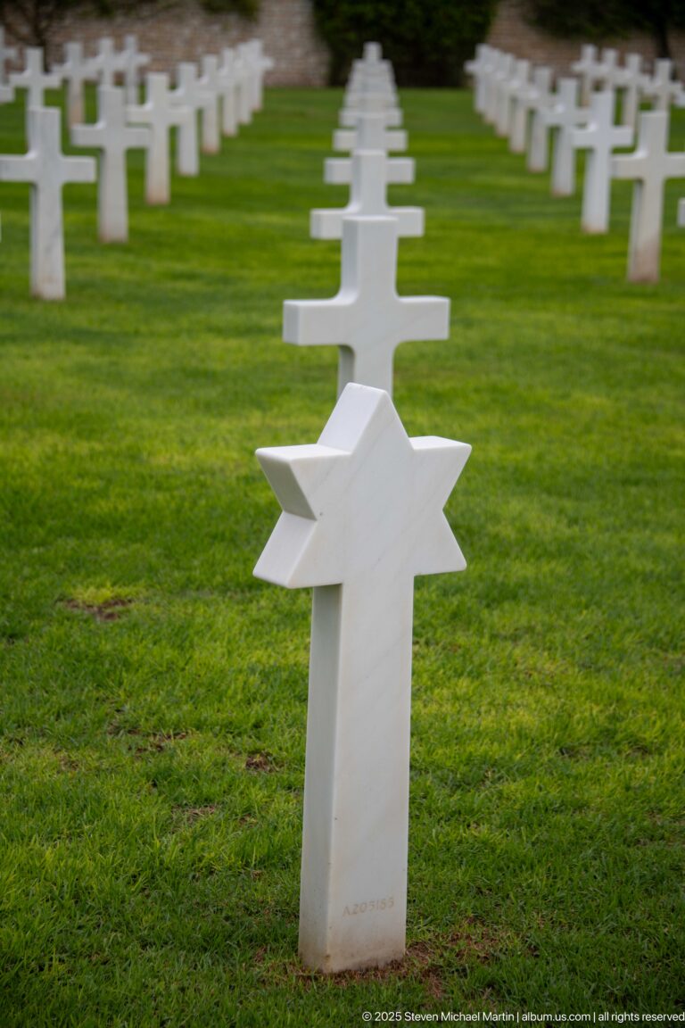 White Star of David grave marker in front of rows of white Latin crosses on grass.
