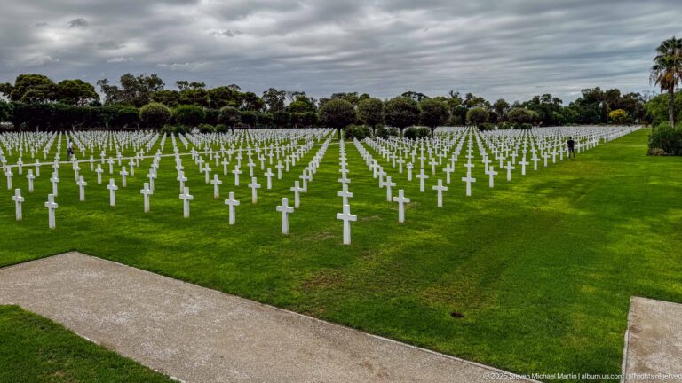 Wide cemetery lawn filled with converging rows of white grave markers beneath dark clouds.