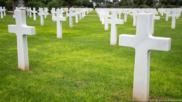 Close white Latin-cross headstone on green grass with rows of matching markers behind.