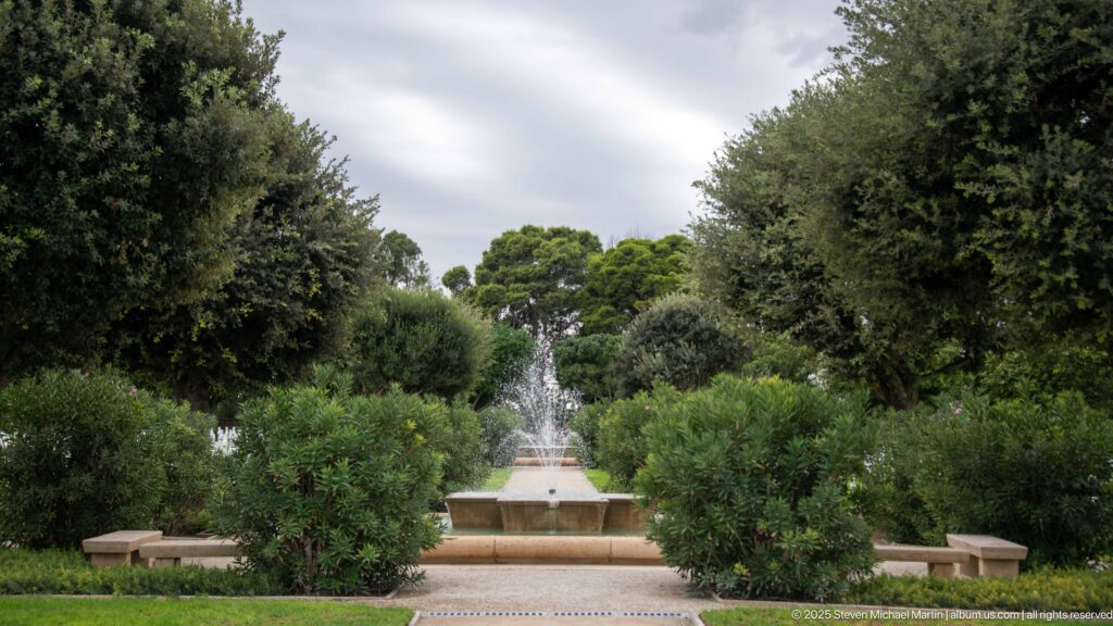 Tiered fountain centered between clipped trees and stone benches along a formal garden axis.