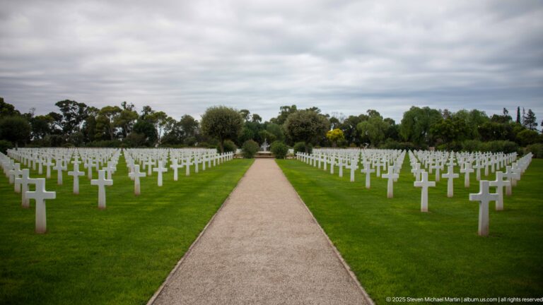Straight pale walkway runs between symmetrical rows of white grave markers toward a distant focal point.