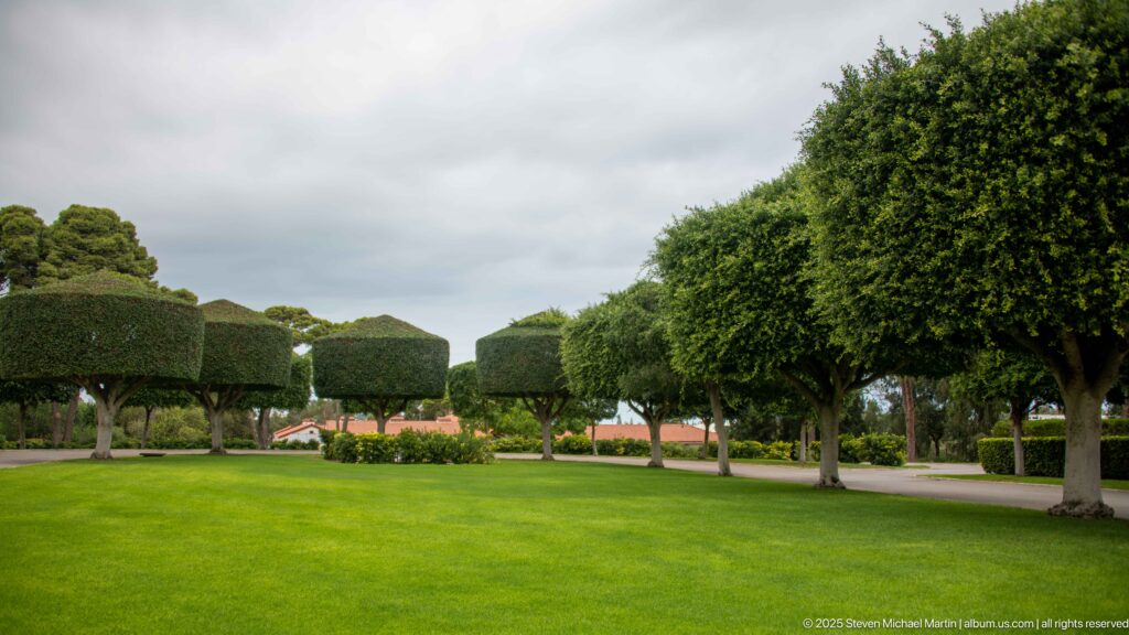 Wide green lawn bordered by clipped trees, hedges, and flowering plantings inside the cemetery.