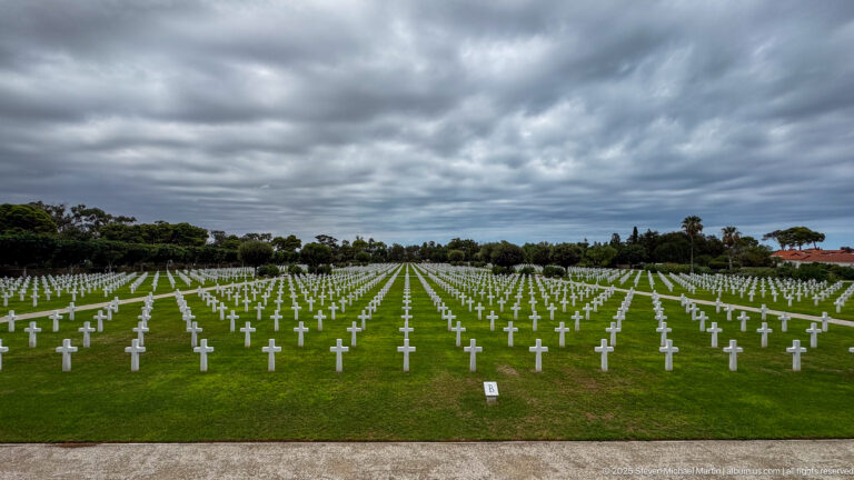 Wide green cemetery lawn filled with rows of white grave markers beneath heavy gray clouds.