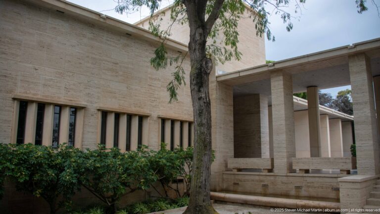 Pale stone memorial building with square columns, narrow windows, steps, and a mature tree in a shaded court.