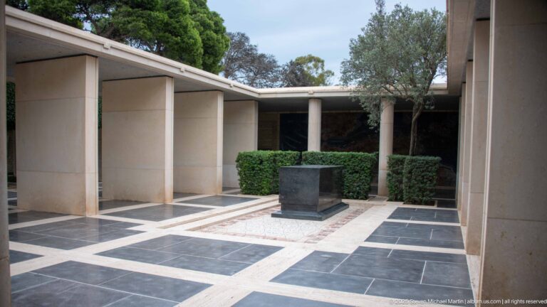 Geometric black-and-pale stone courtyard with a dark rectangular pedestal, columns, clipped hedges, and a tree.