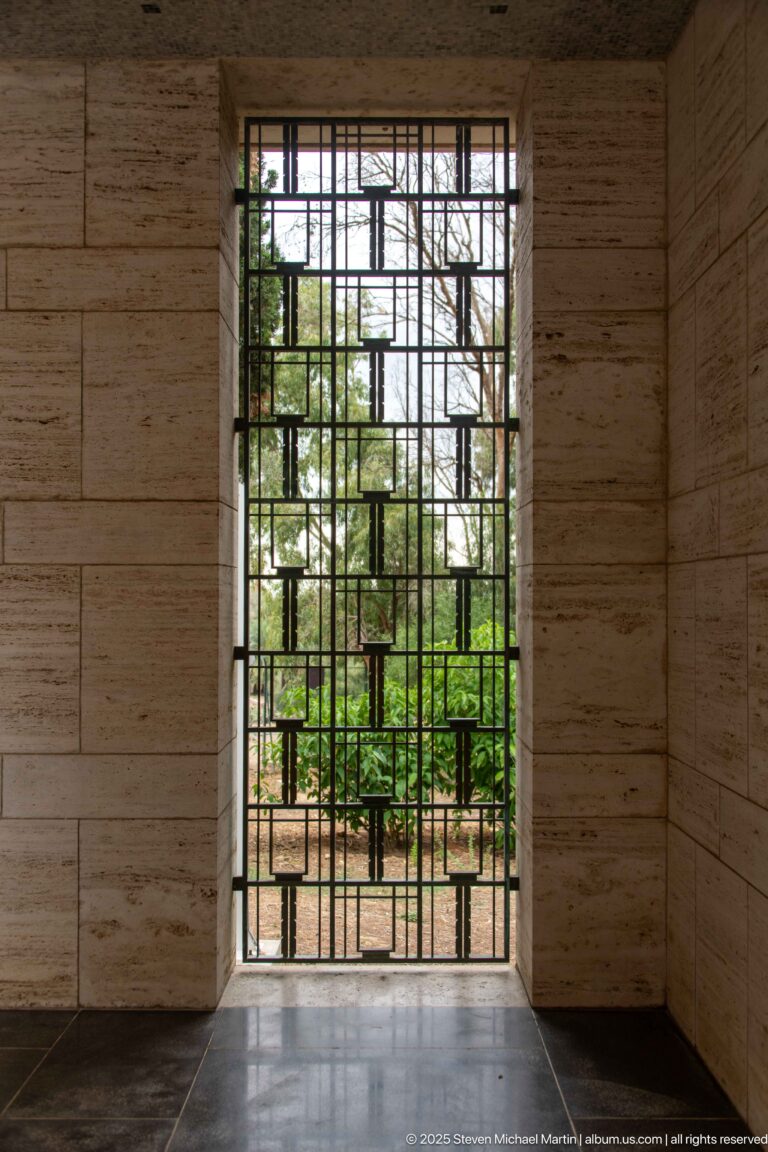 Dark geometric metal grille in a pale stone opening with green cemetery landscaping visible beyond.