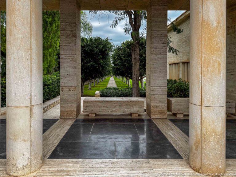 View between pale stone columns toward a long straight path lined with clipped trees and hedges.
