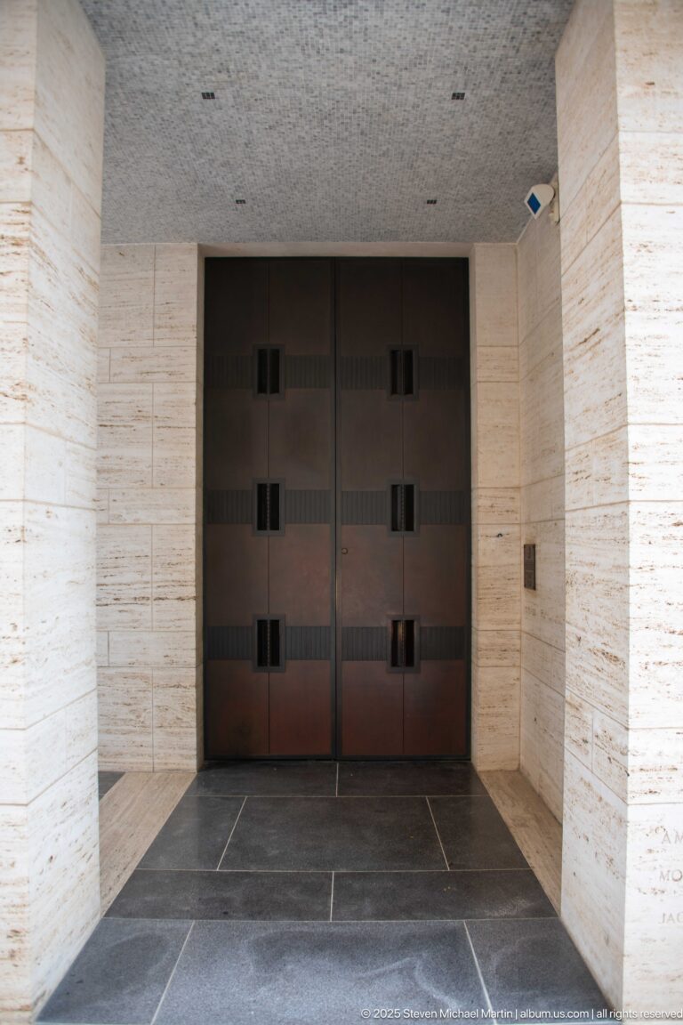 Large dark double doors with rectangular inset panels at the end of a pale stone vestibule.
