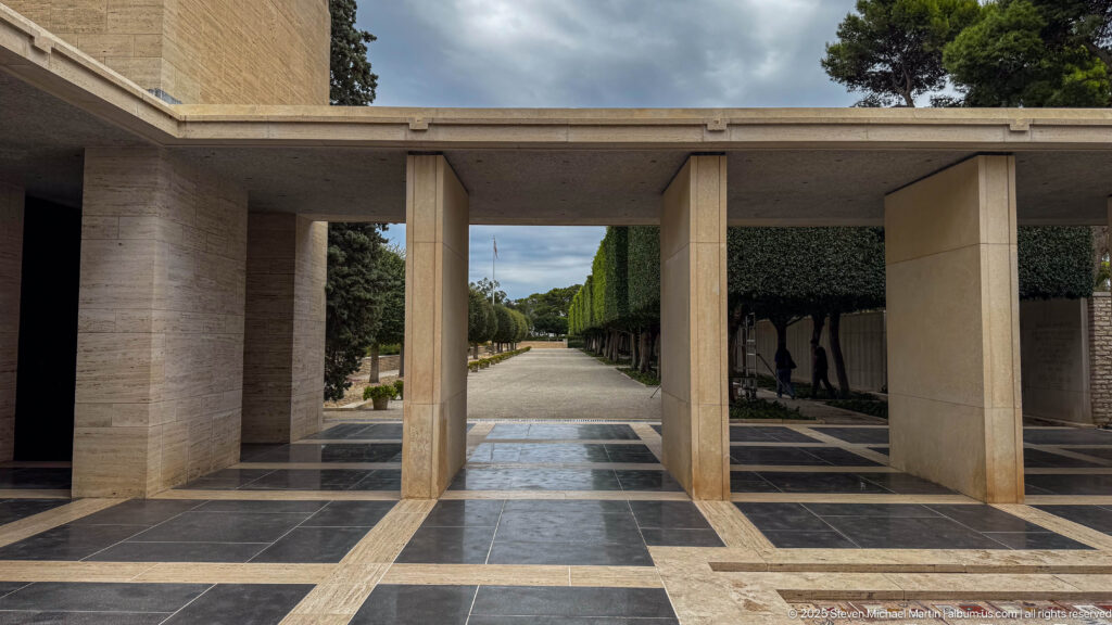 Open pale stone portico with dark floor paving looking toward a straight tree-lined cemetery path.