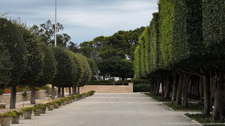 Broad gravel path bordered by tightly clipped rows of trees and hedges in the cemetery grounds.