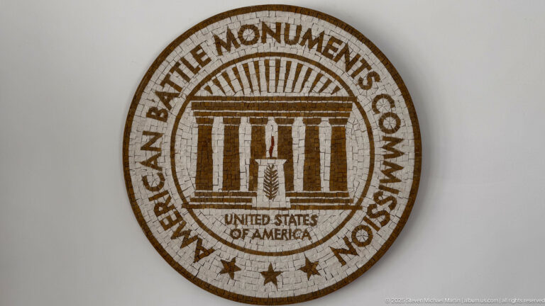 Circular American Battle Monuments Commission seal with a classical colonnade motif and surrounding lettering.