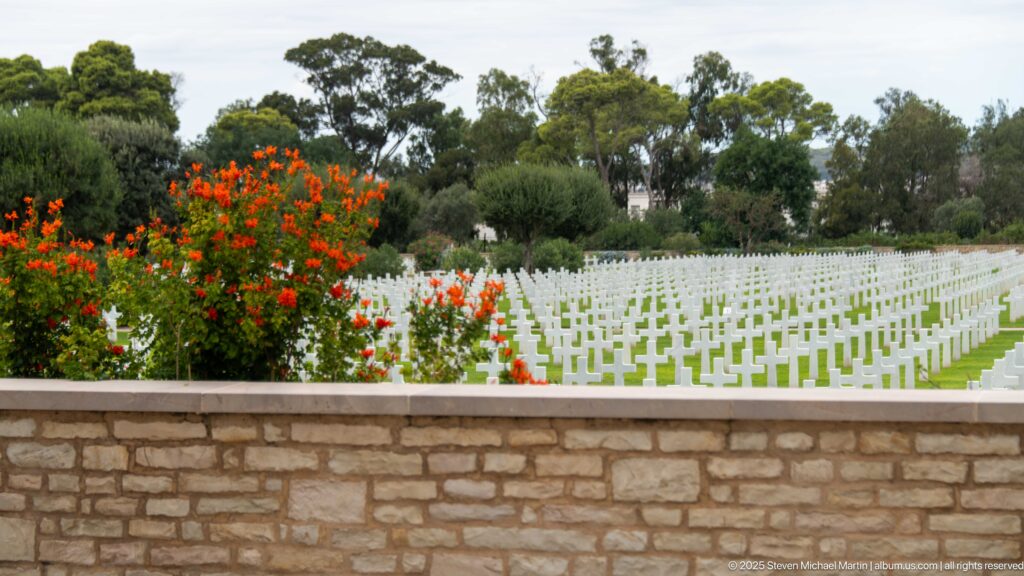 White grave markers across a green lawn beyond red flowers and a low tan stone wall.