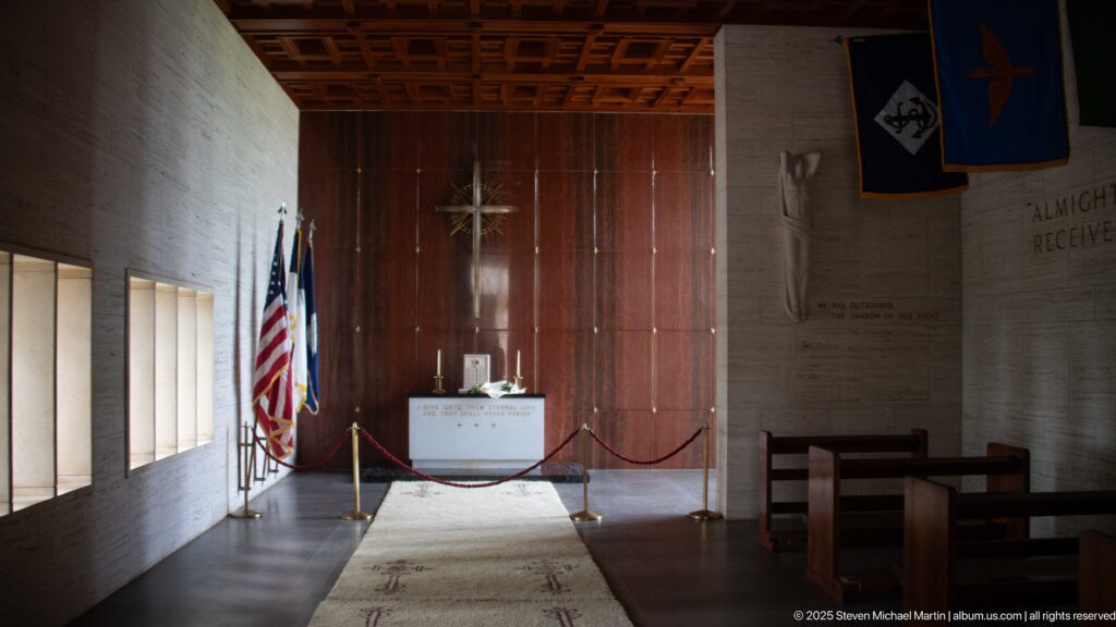 Memorial chapel interior with altar, large cross, flags, military banners, pews, and a central carpet runner.