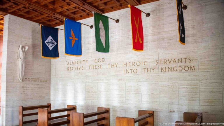 Military service banners hang above a chapel wall inscribed Almighty God receive these thy heroic servants into thy kingdom.