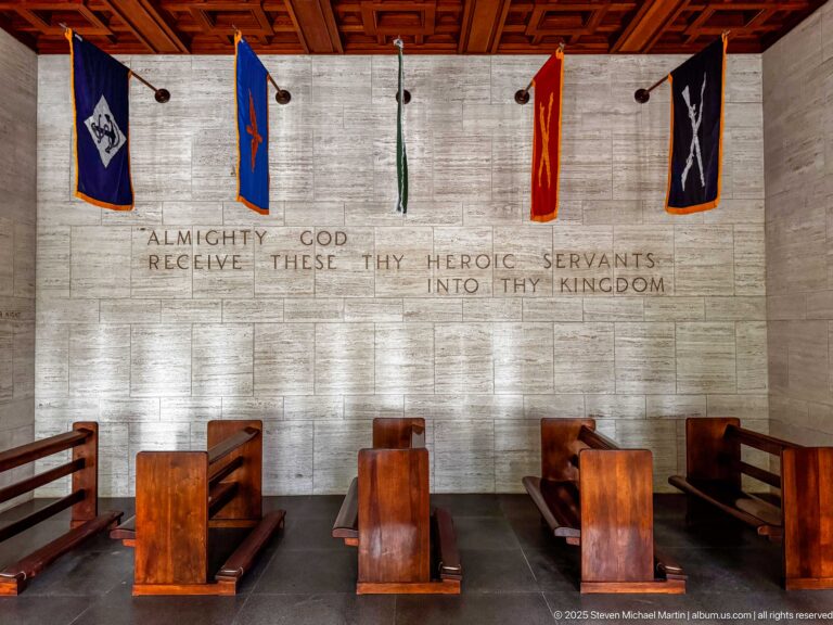 Rows of wooden pews face a stone chapel wall with military banners and a carved devotional inscription.
