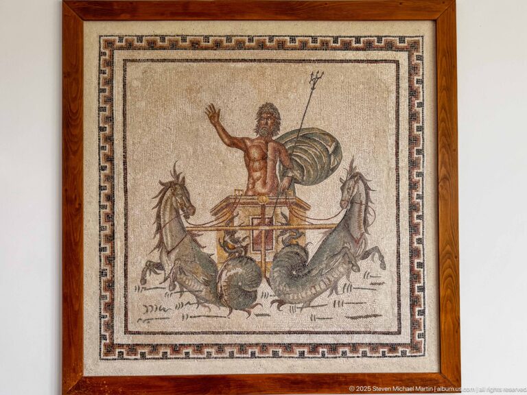Framed classical-style mosaic showing a trident-bearing sea god in a chariot drawn by marine horses.