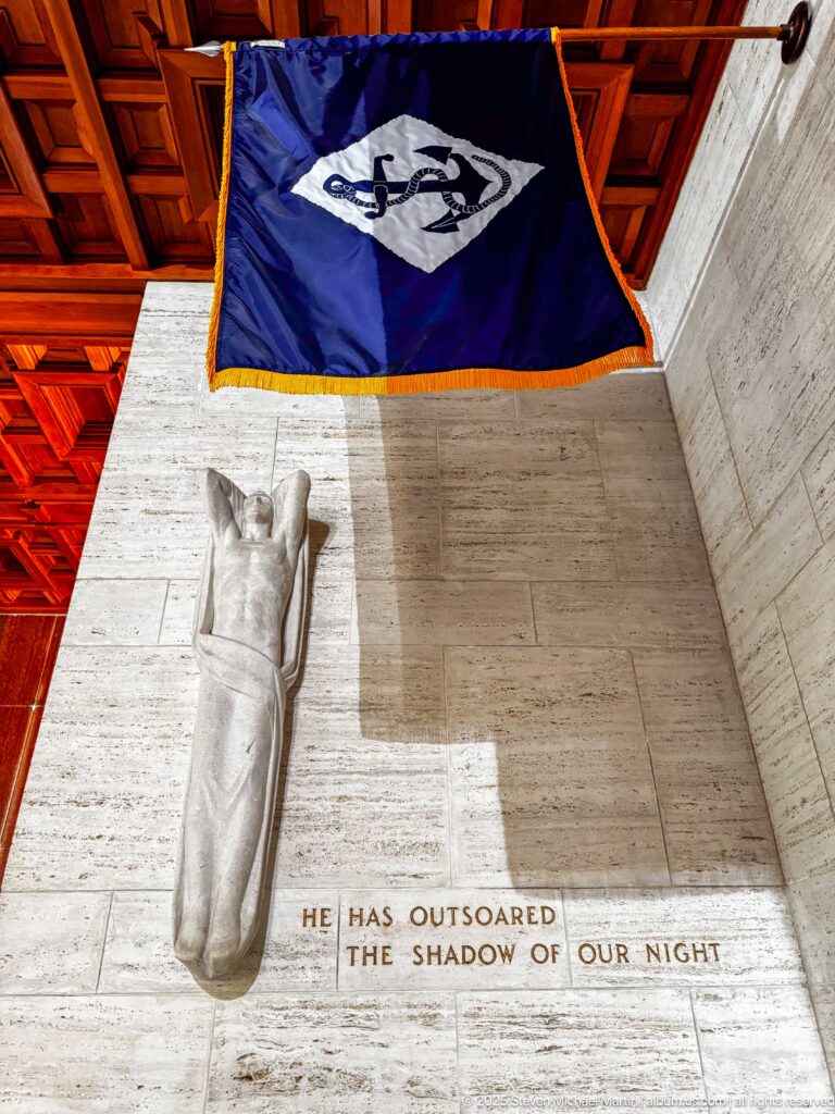 Draped relief figure on a pale stone chapel wall beneath a blue banner and the inscription He has outsoared the shadow of our night.