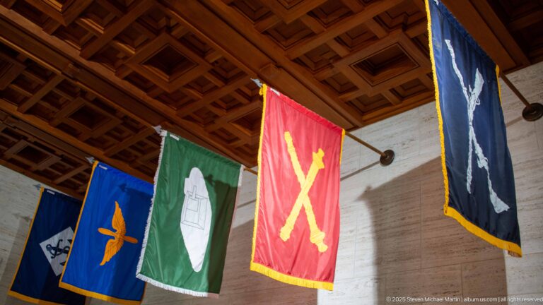 Blue, green, red, and dark military service banners hang beneath a geometric wooden chapel ceiling.