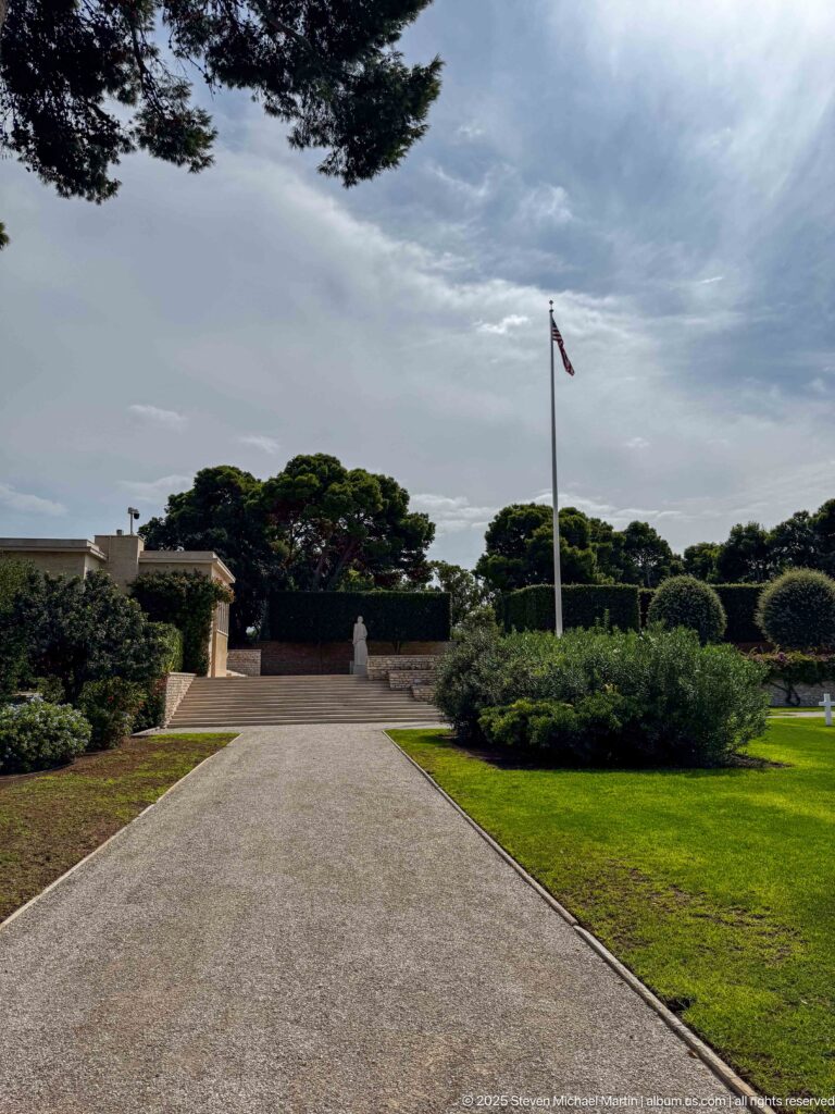 Straight gravel path bordered by clipped shrubs leading toward a tall American flagpole.
